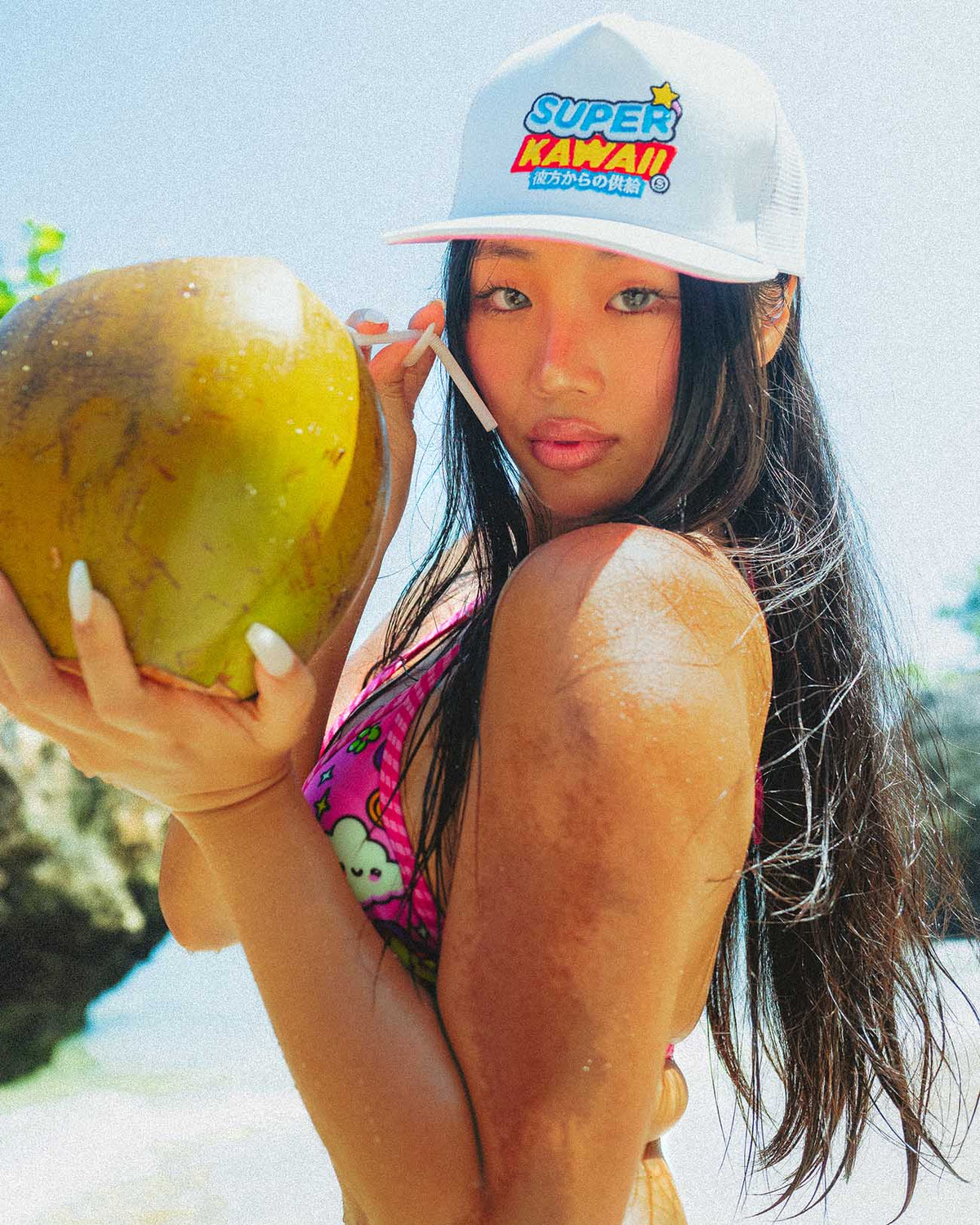 Model in pink bikini holding coconut, wearing SUPERS KAWAII hat on beach, showcasing summer fashion and playful vibes.