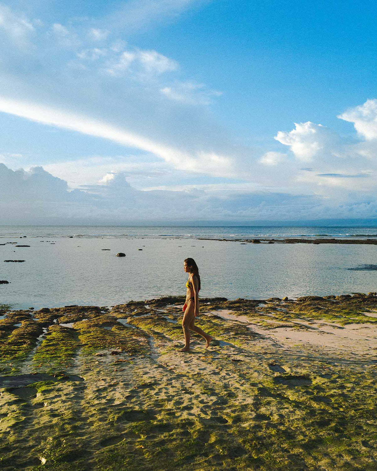 Model wearing Samba Sunrise - (Thong Set) bikini walking on beach under blue sky.