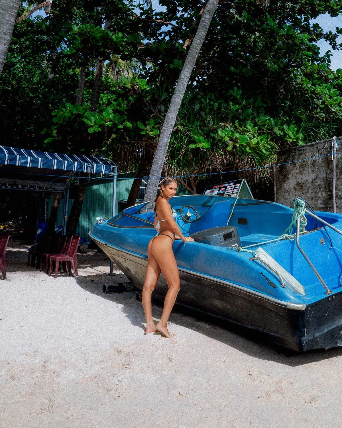 Adjustable white triangle bikini top and green thong bottom on a model, standing next to a blue boat on a beach.