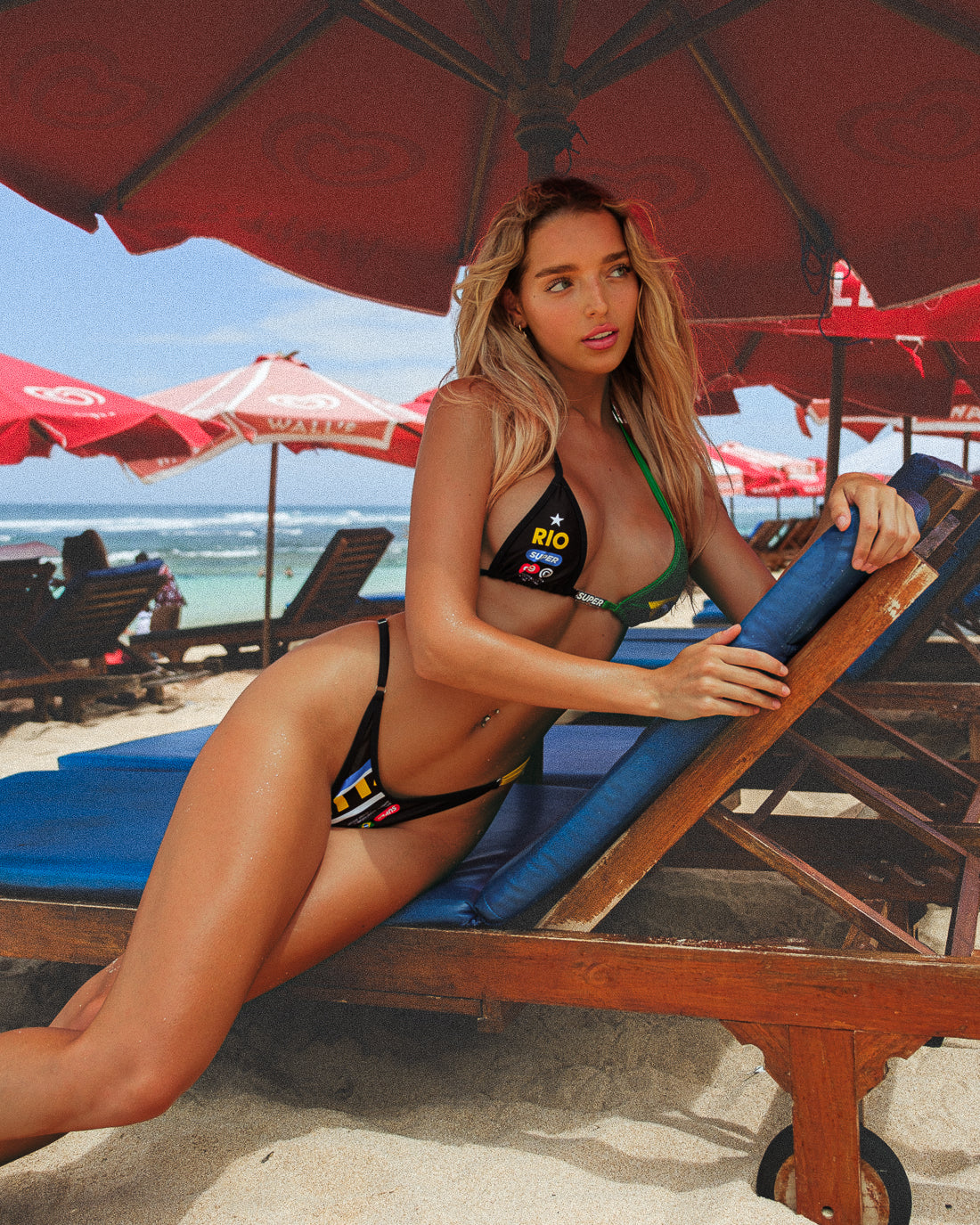 In a close-up side body shot, the model sits on a beach bench posing in a sexy, black women's bikini with Rio-inspired print. The classic triangle top is embellished with a cute charm bar, paired with high-waisted, adjustable-side thong bottoms, creating a cute and comfortable beach look. - Alternate view